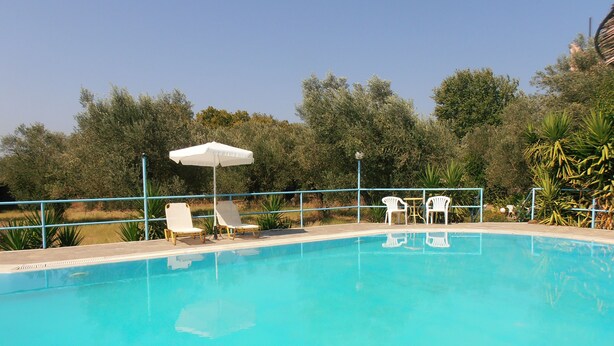 A swimming pool with white lounge chairs and a white umbrella. - Kipos Studios (Lesvos)