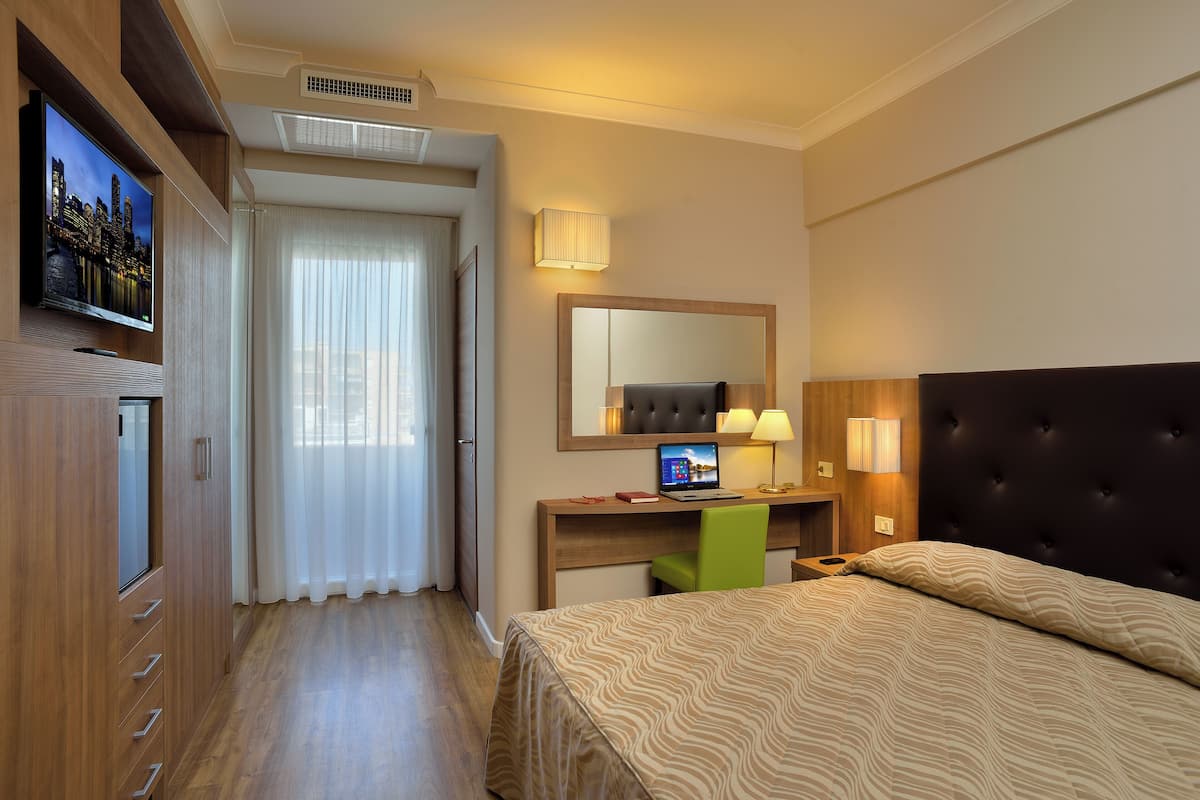 Habitación de hotel con una cama, un escritorio con una computadora portátil, una silla verde, un televisor montado en la pared y un amplio ventanal con cortinas.