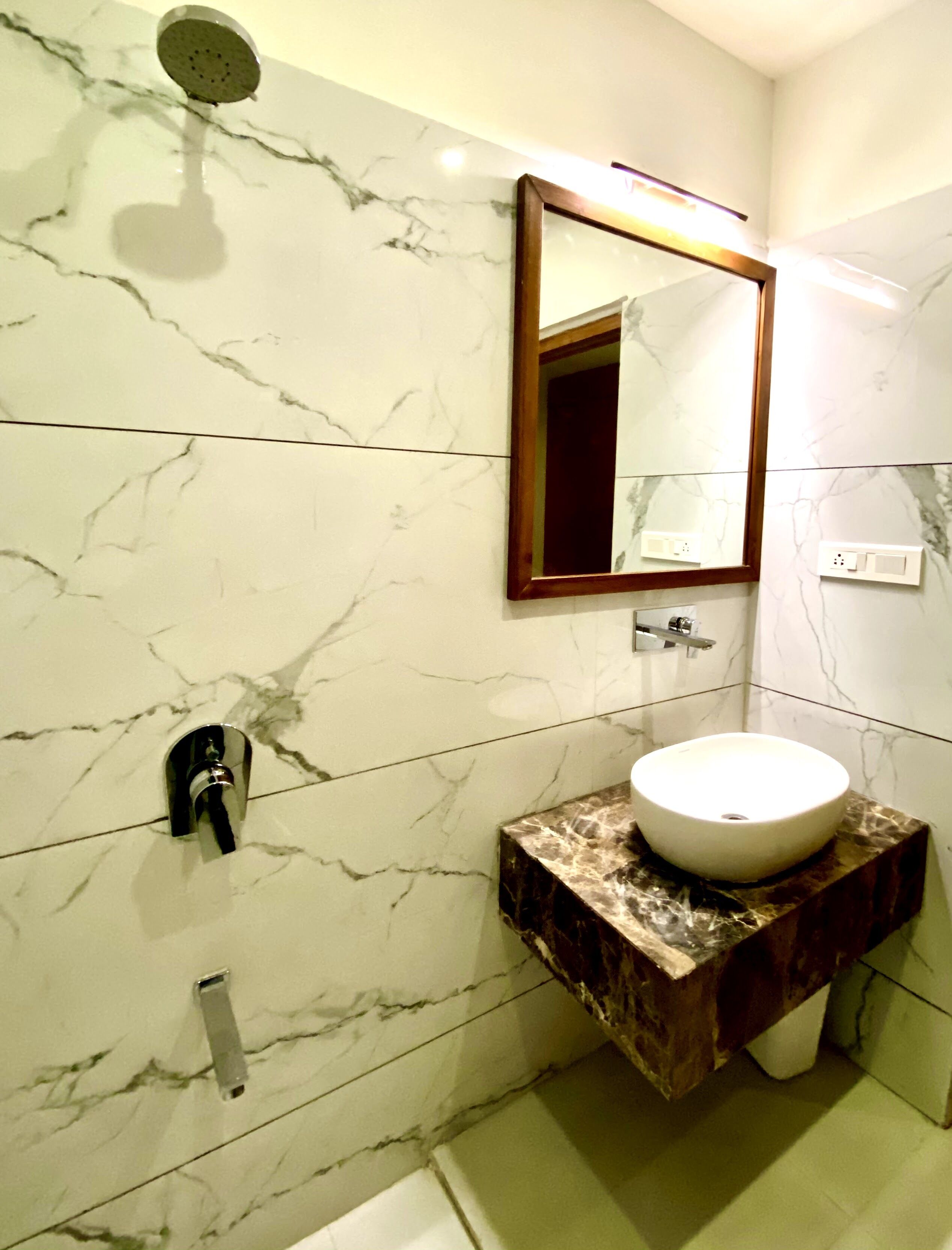 Un baño con pared de mármol, un lavamanos con encimera de mármol, un espejo y un cabezal de ducha.