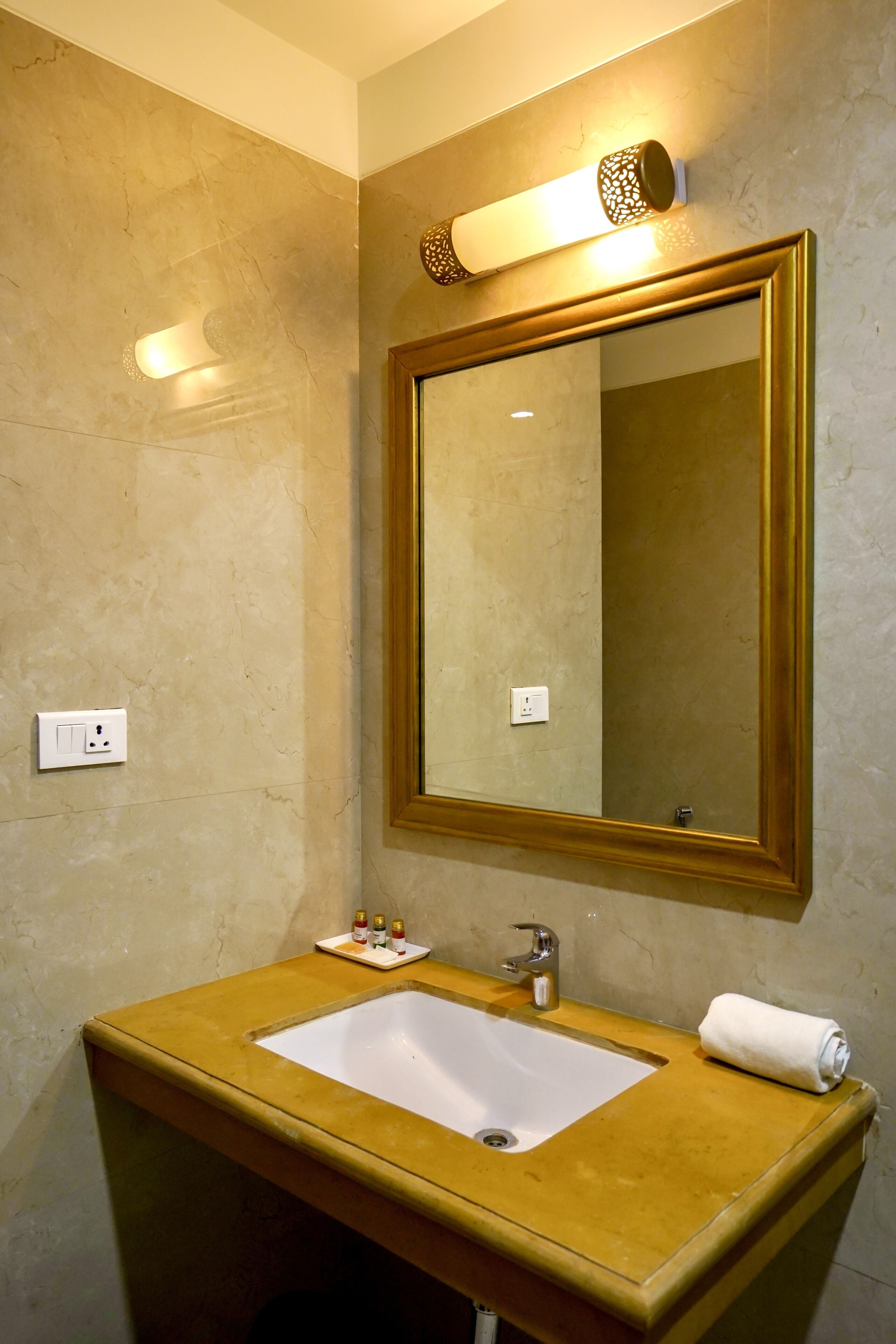 A bathroom with a beige marble wall, a gold-framed mirror, a white sink, and a towel on the counter.