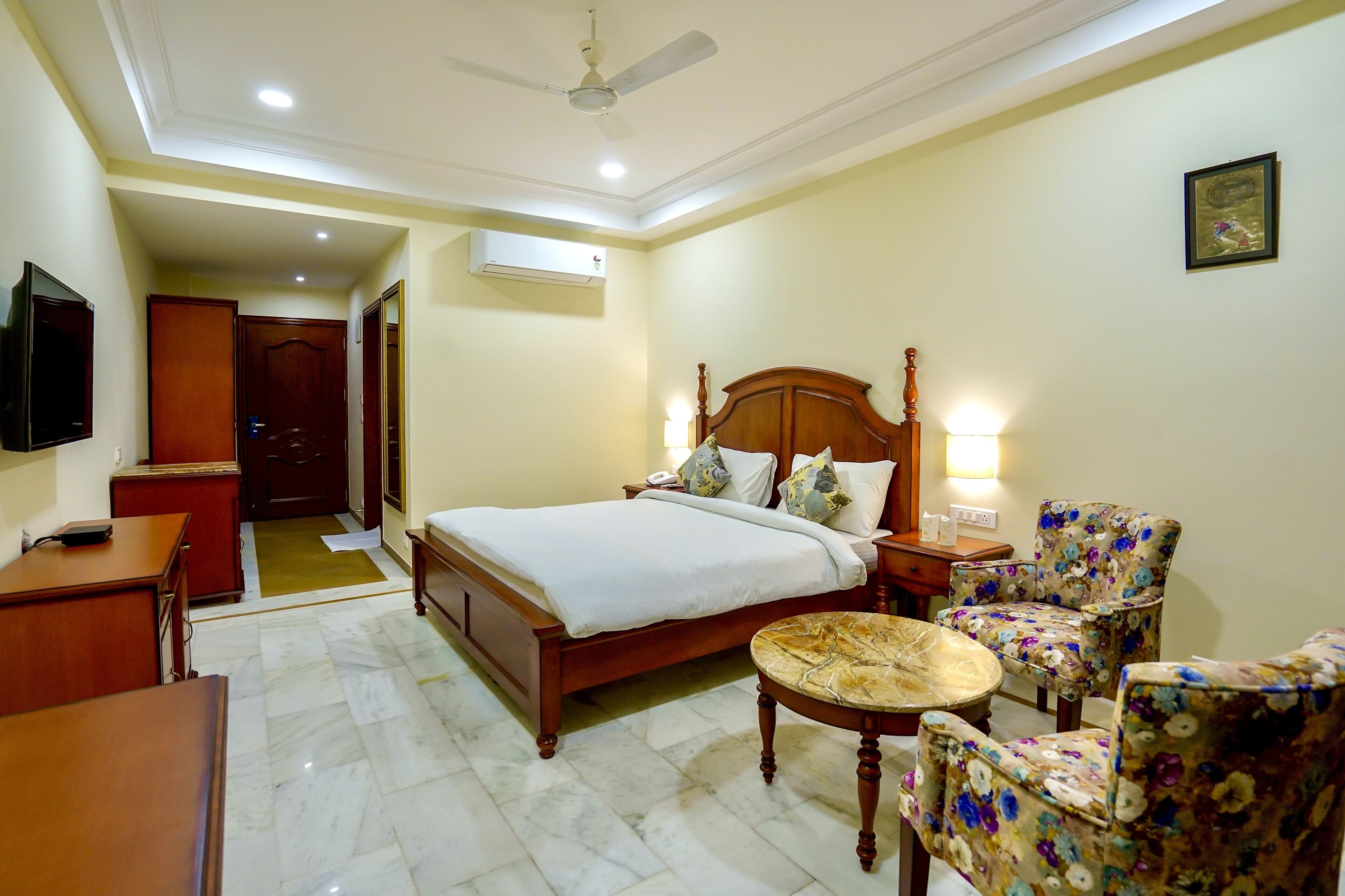 A hotel room with a bed, a desk, a chair, a round table, a ceiling fan, and a TV.