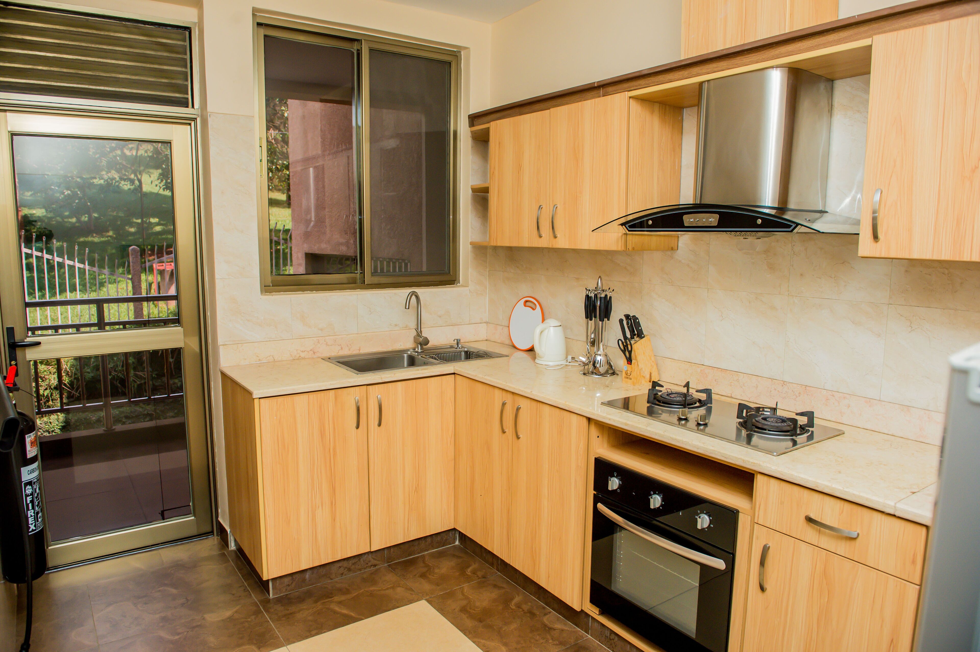 A modern kitchen with wooden cabinets, a stainless steel range hood, a dishwasher, and a window with a view of a garden.