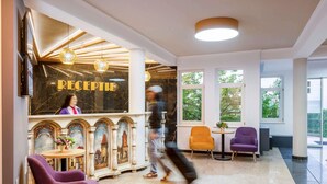 Un área de recepción con pared de mármol, mostrador de recepción y un amplio ventanal con vista a árboles. Hay dos sillones y una mesa pequeña con un jarrón de flores.
