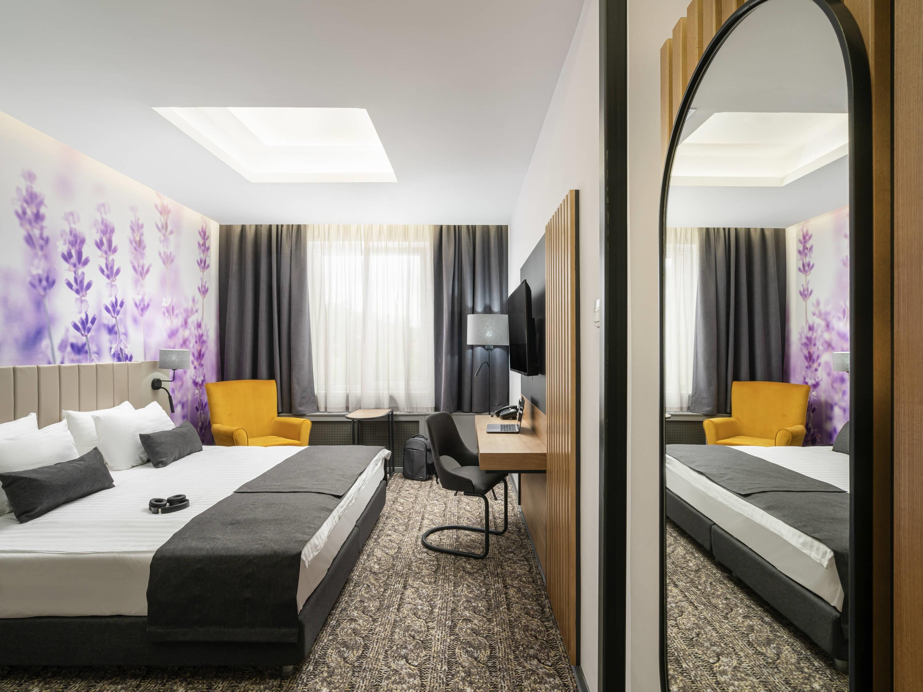 A hotel room with a bed, a desk, a chair, a TV, and a large mirror.