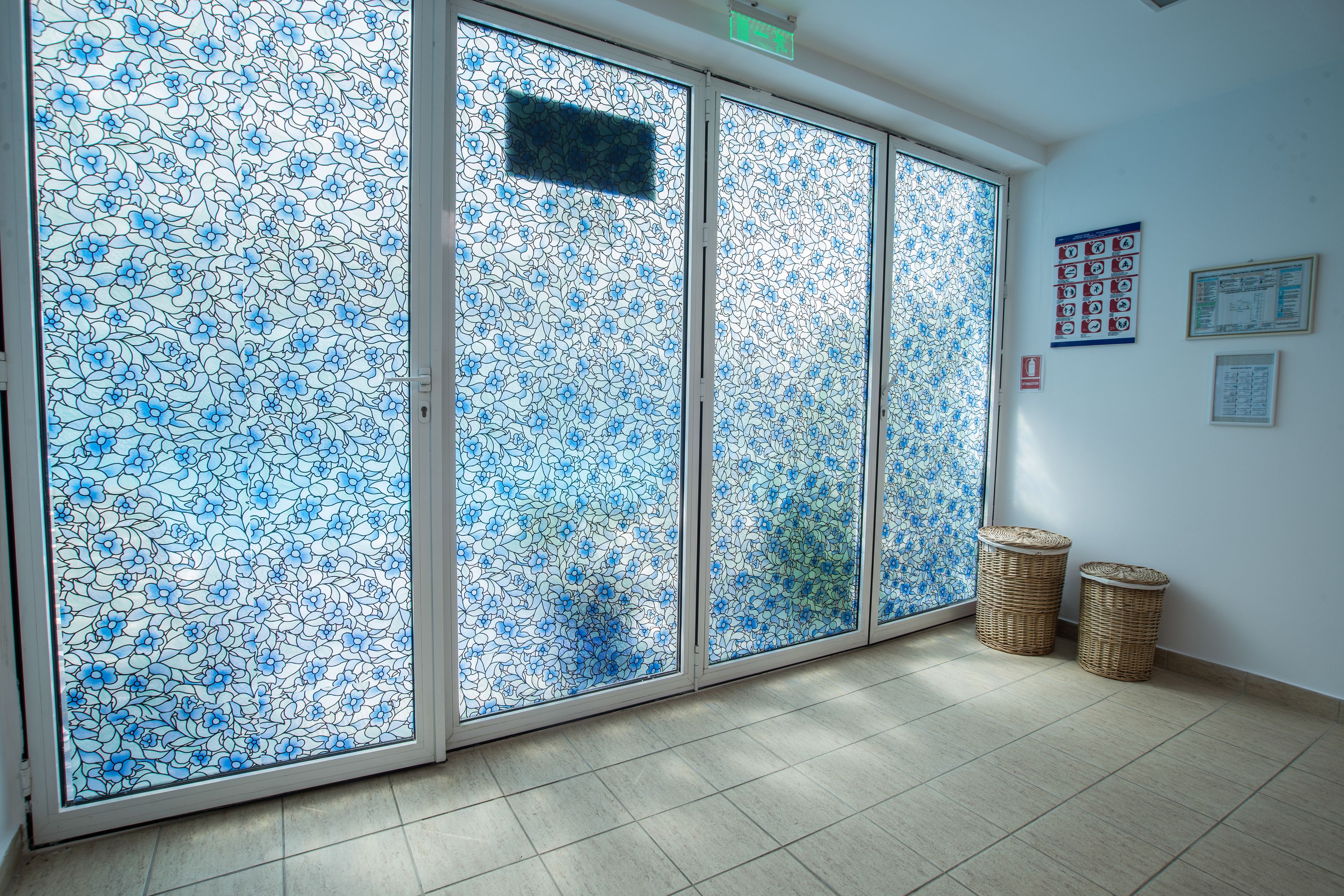 Una habitación con un amplio ventanal corredero de diseño floral azul, dos cestas de mimbre en el suelo y una pared con carteles informativos.