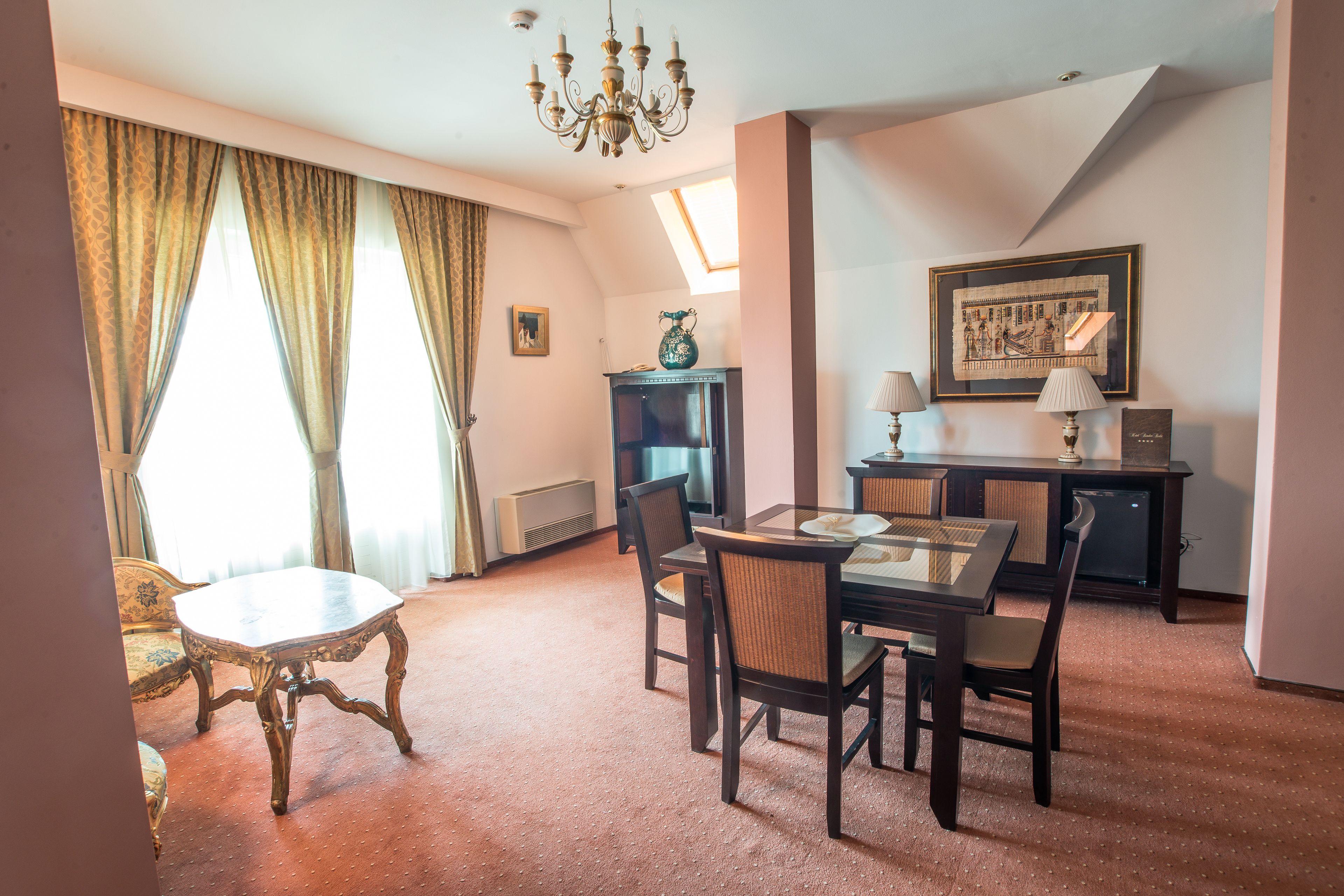 A hotel room with a dining table, chairs, a chandelier, a painting, and a window with curtains.