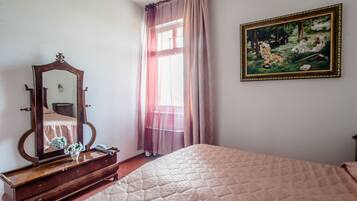 Un dormitorio con una cama, un aparador, un espejo, un cuadro y una ventana con cortinas.