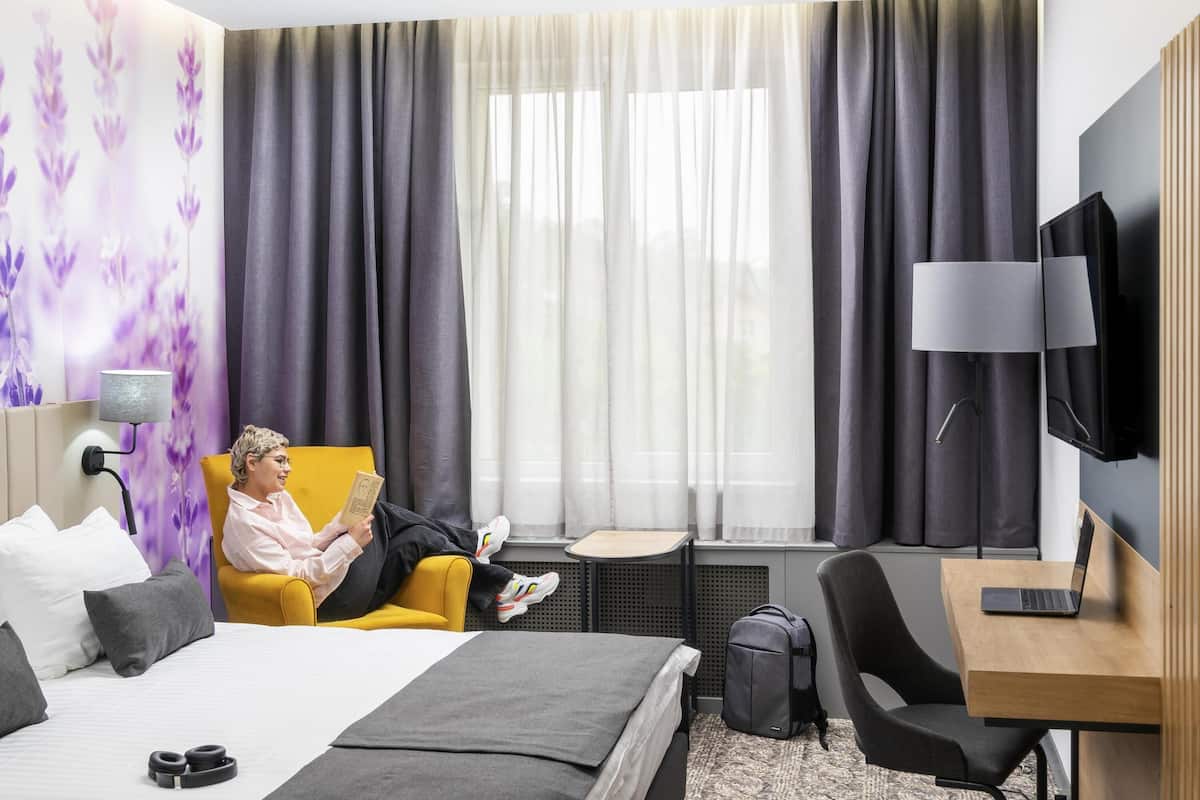 A hotel room with a bed, a desk, a chair, a TV, and a window with curtains.