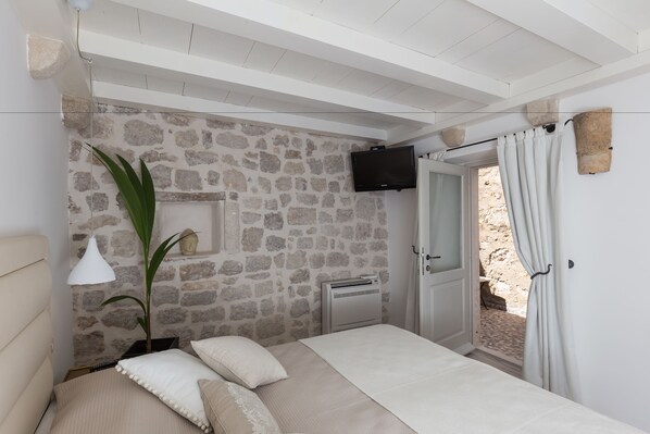 A bedroom with a stone wall, a bed with white bedding, a TV mounted on the wall, and a door leading to an outdoor area. - Dubrovnik Sweet House (Dubrovnik)