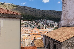 A town with terracotta-roofed buildings, a hillside, and a prominent stone wall.