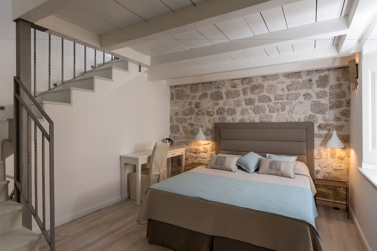 A bedroom with a stone wall, a bed with a headboard, a desk, and a staircase.