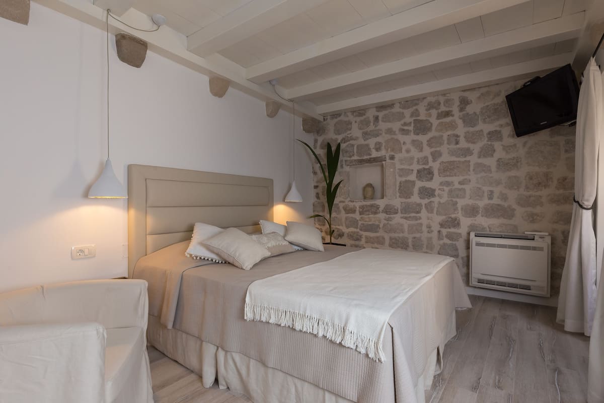 A bedroom with a stone wall, a bed with a headboard, a bedside table, a lamp, and a wall-mounted TV.