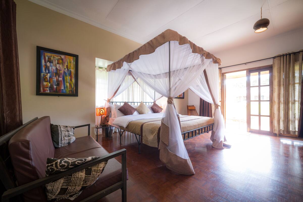 A bedroom with a four-poster bed, a sofa, a painting on the wall, and a window with curtains.