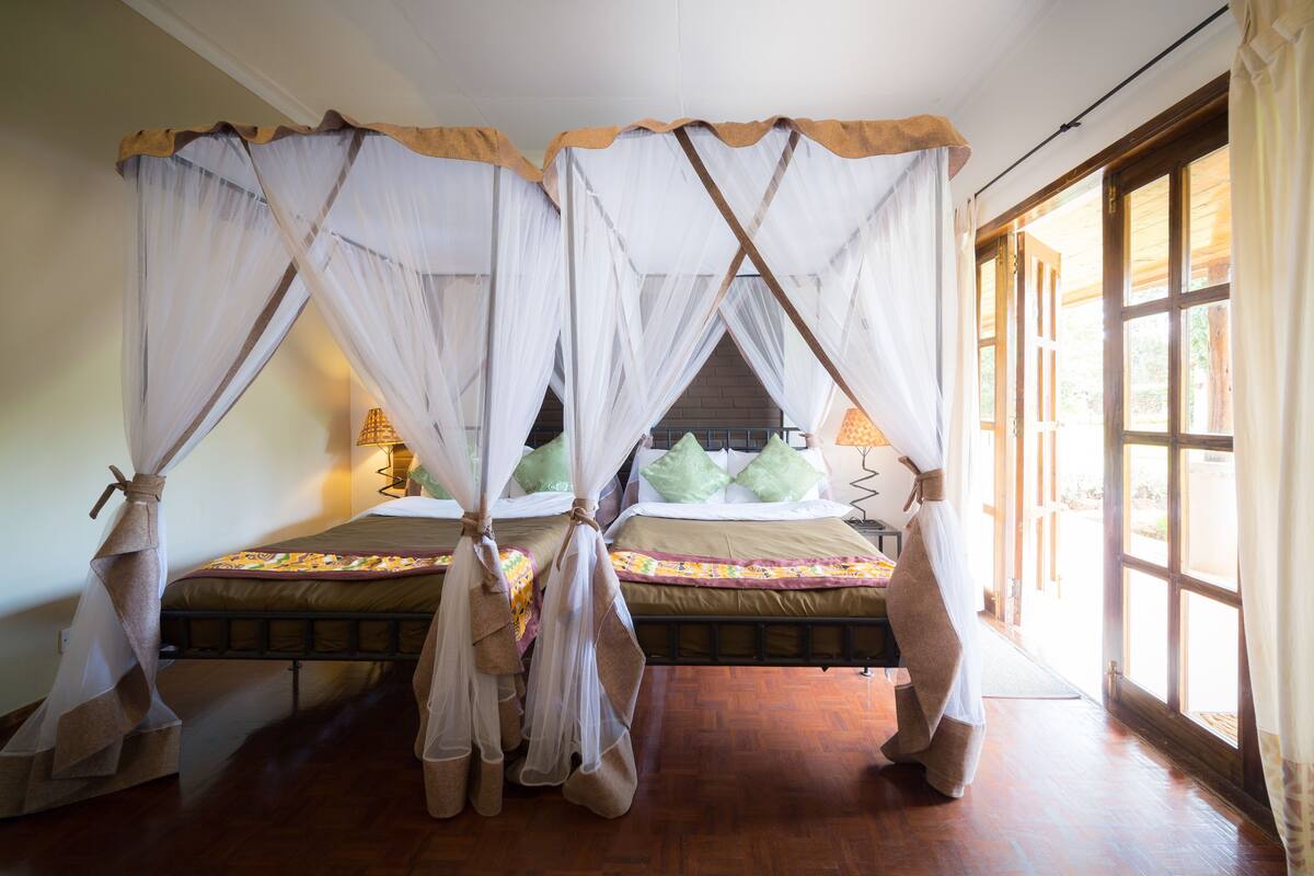 A room with two beds, each with a canopy bedspread and pillows, a wooden floor, and a large window with curtains.