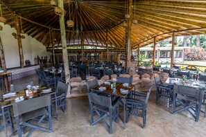 A spacious outdoor dining area with multiple tables and chairs, a thatched roof, and a view of the surrounding landscape.