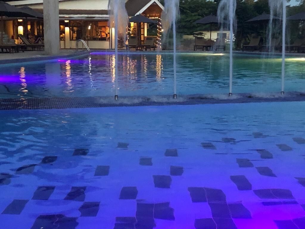 A swimming pool with tiled flooring, a lounge area with chairs and umbrellas, and a building with lights in the background.