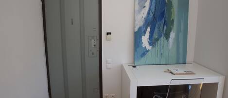 A room with a white desk, a clear chair, a glass door, and a painting on the wall.