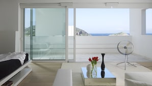 Un salon moderne avec une porte vitrée donnant sur un balcon, un canapé blanc, une table basse ornée d’un vase de fleurs rouges et un ventilateur sur pied.