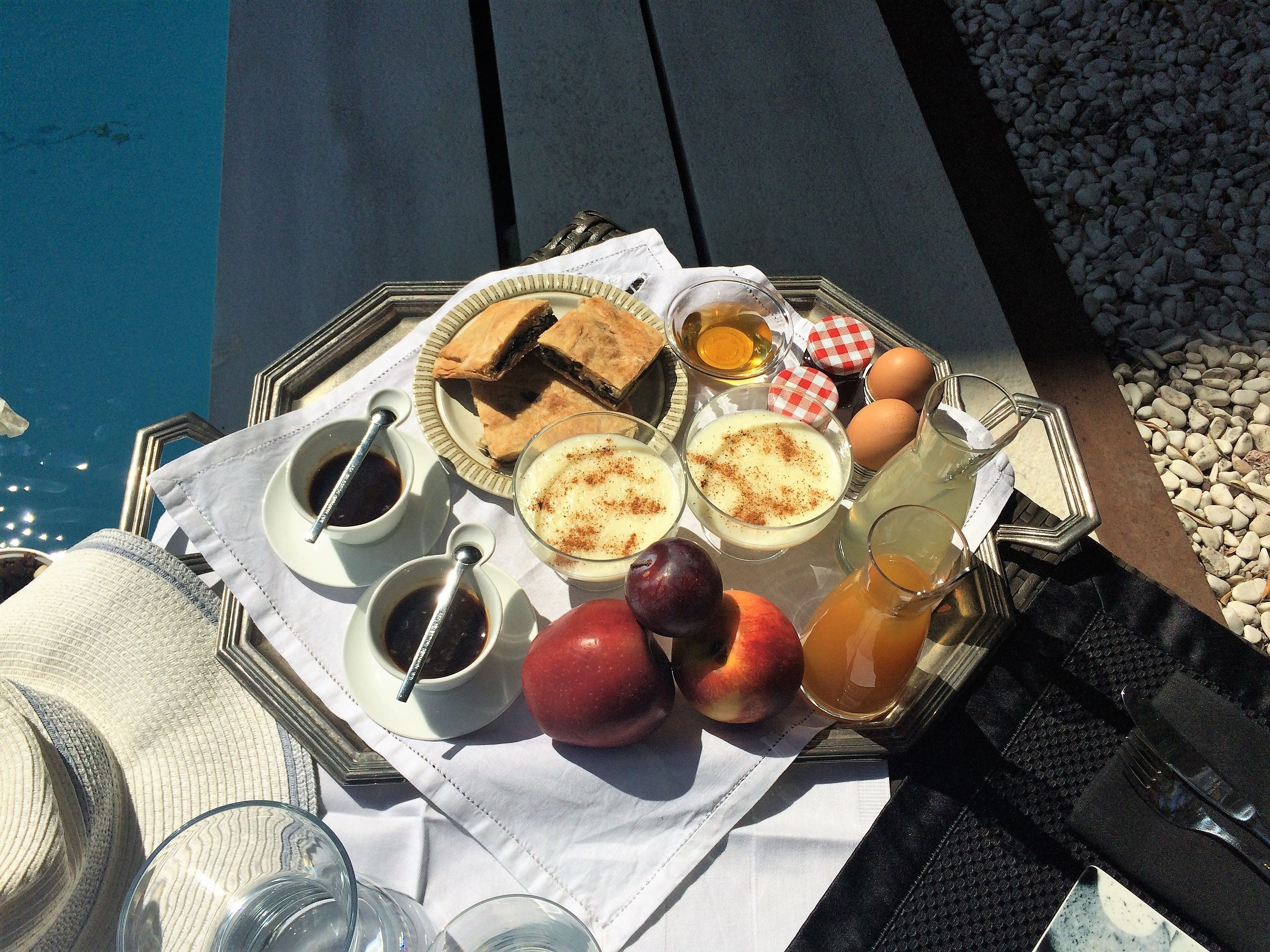 Un plateau de petit-déjeuner avec des viennoiseries, des œufs, des fruits et des boissons, servi au bord de la piscine.