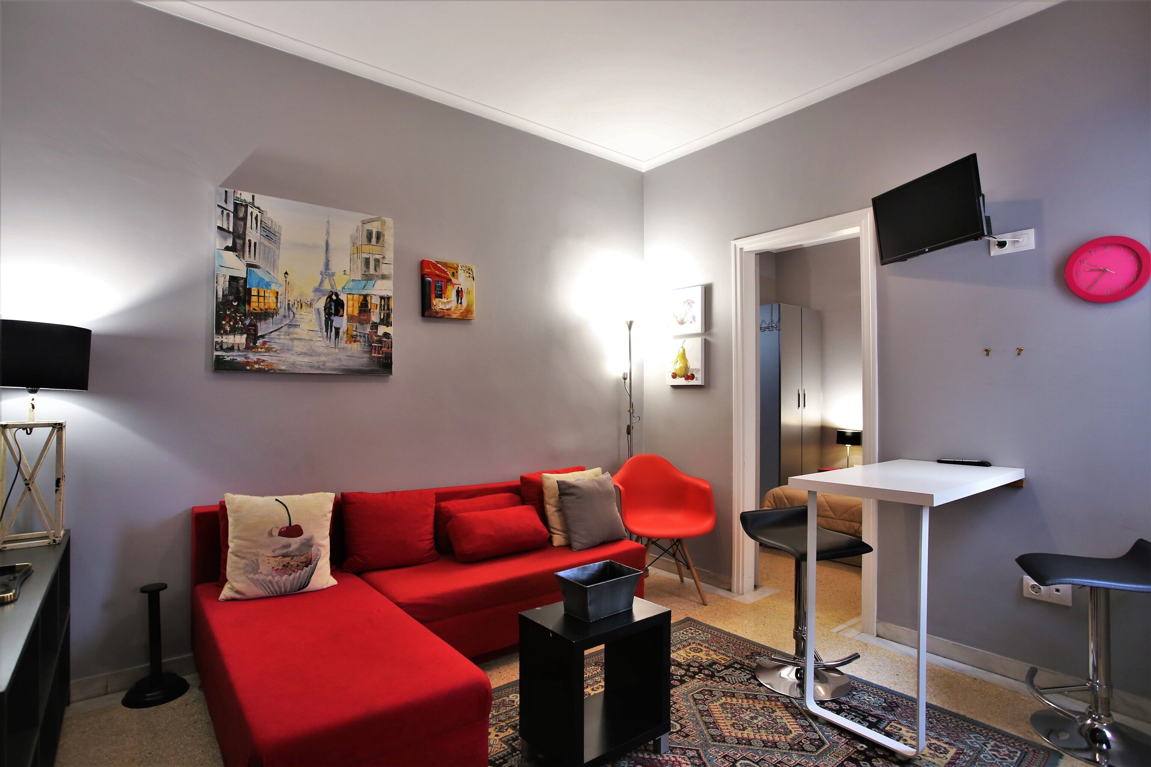 Un salon moderne avec un canapé rouge, une petite table blanche, un téléviseur fixé au mur et une horloge rose.