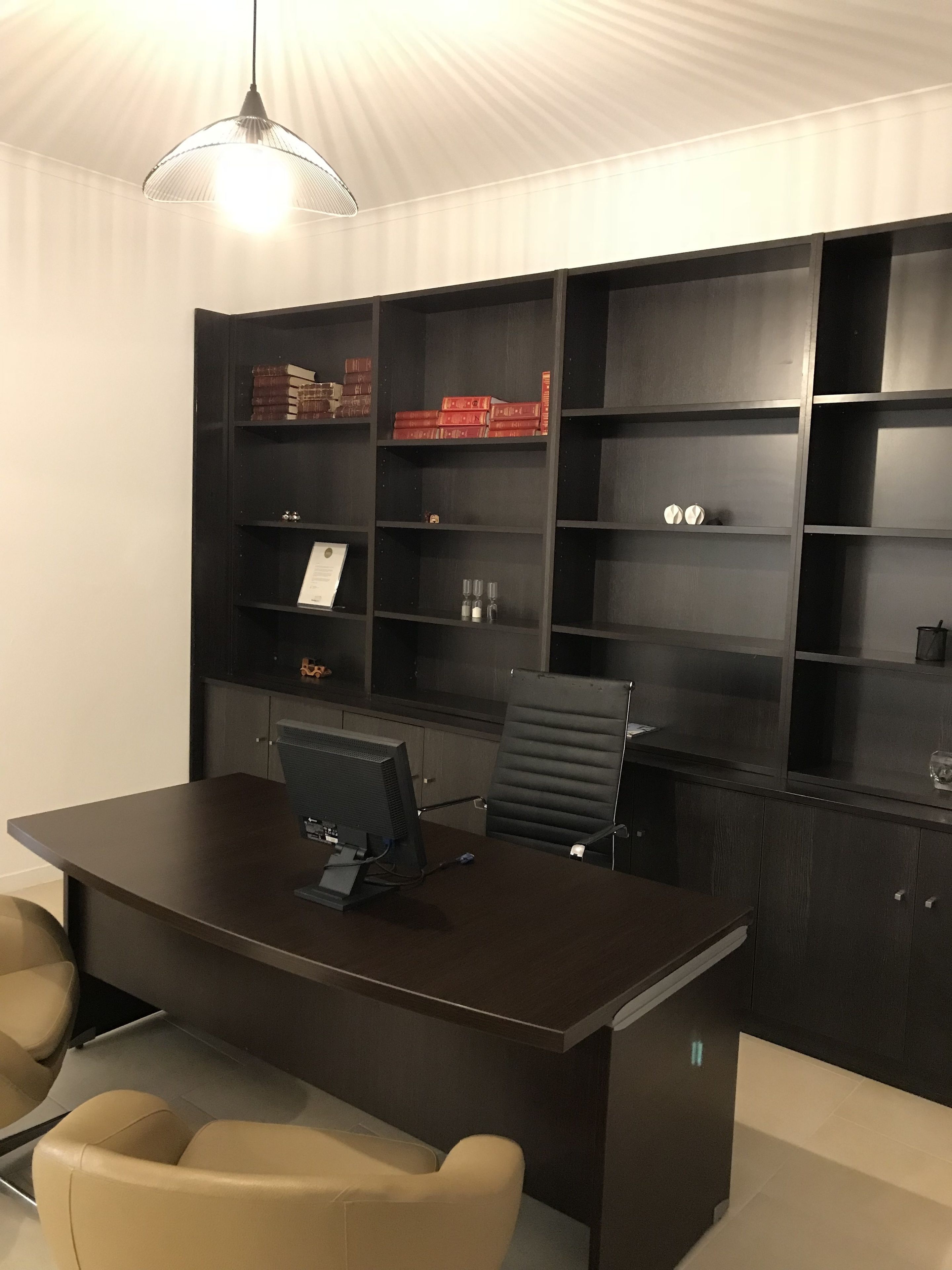 Een modern kantoor met een donker houten bureau, een computer en een zwarte stoel. Er staan boekenkasten met boeken en decoratieve objecten.