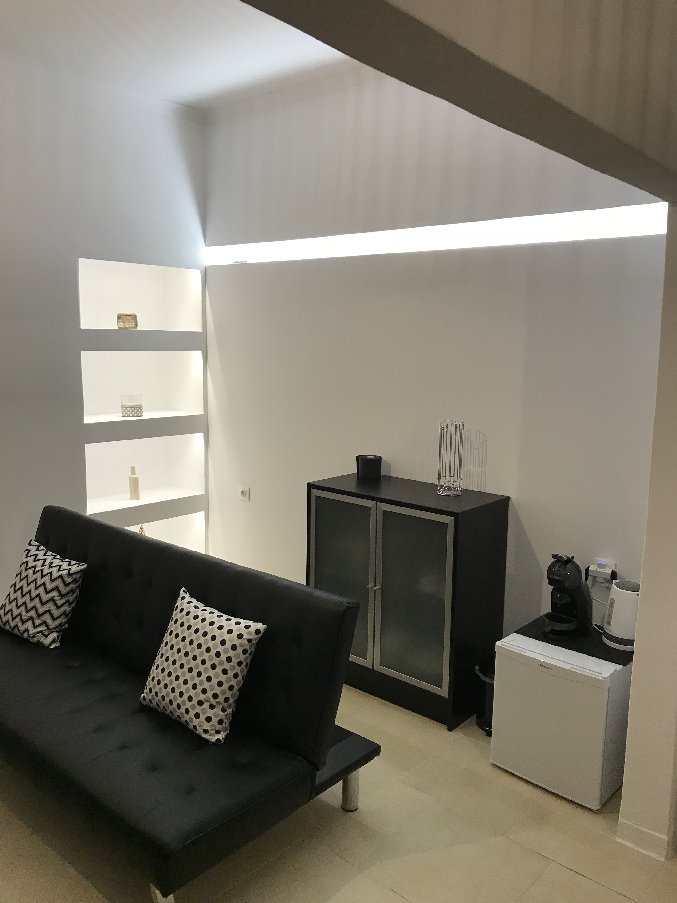 A modern living room with a black sofa, two patterned pillows, a cabinet with a coffee machine, and a small refrigerator.
