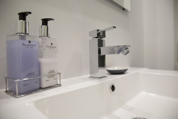 A bathroom with a white sink, two bottles of Pecksniff's body lotion, and a modern faucet.