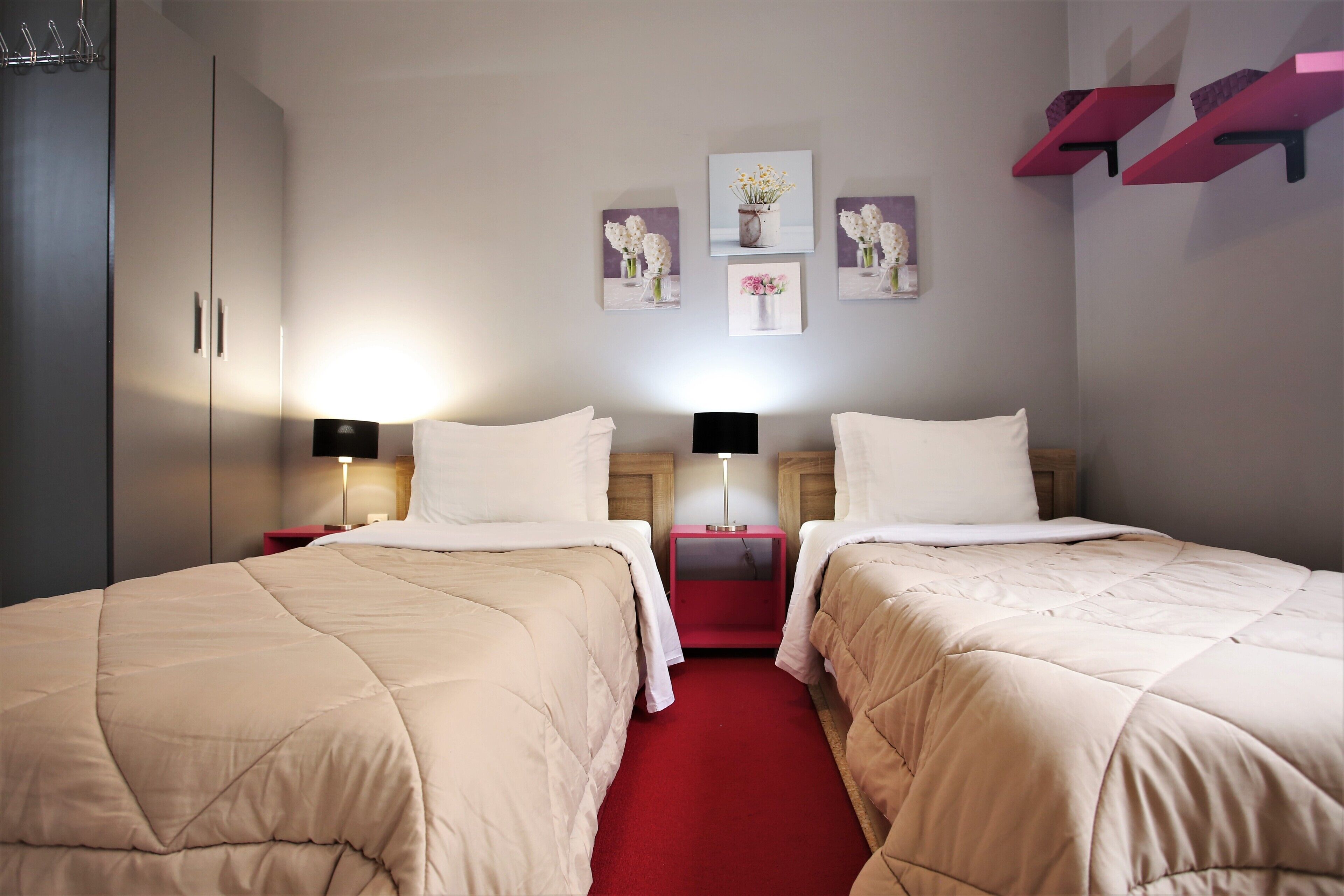 A hotel room with two beds, a red carpet, a wardrobe, and wall-mounted artwork.