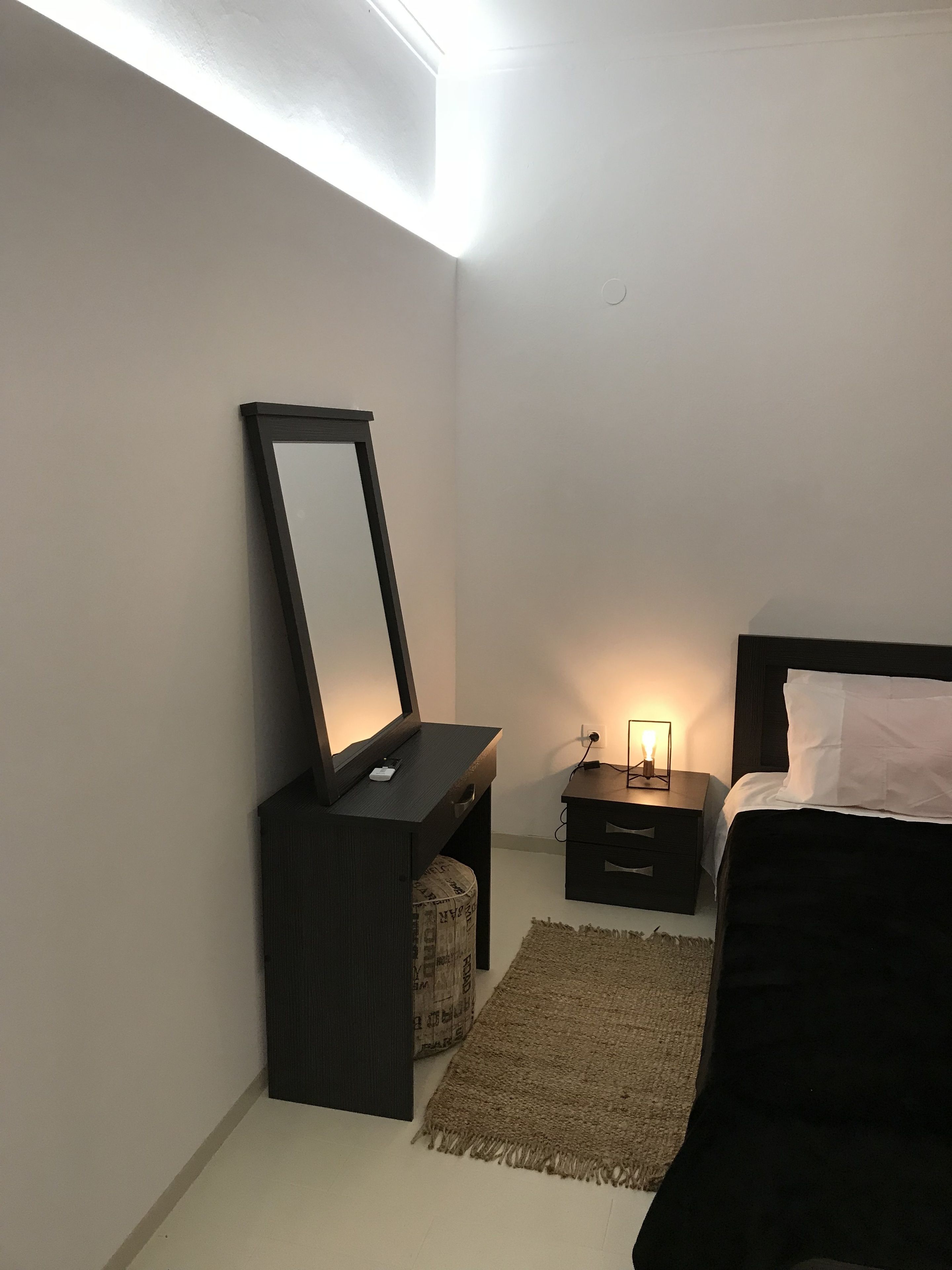 Un dormitorio con cama, mesita de noche con lámpara, cómoda con espejo y alfombra.