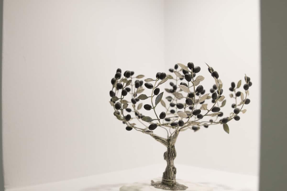 A sculpture of a tree with a square base on a white pedestal in a room with white walls and a white floor.