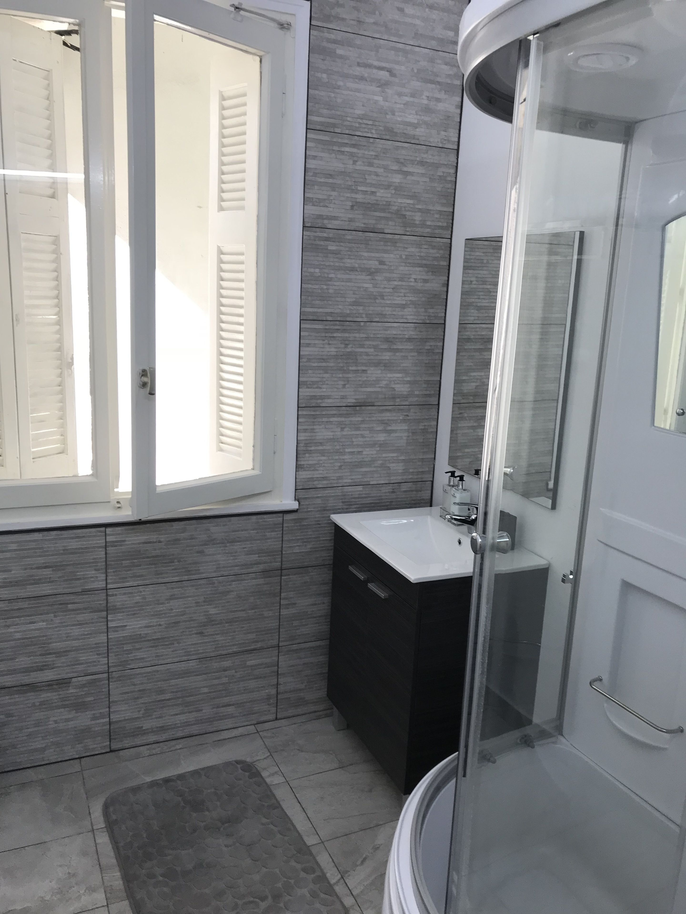 A modern bathroom with a glass shower enclosure, a white sink, and a window with white shutters.