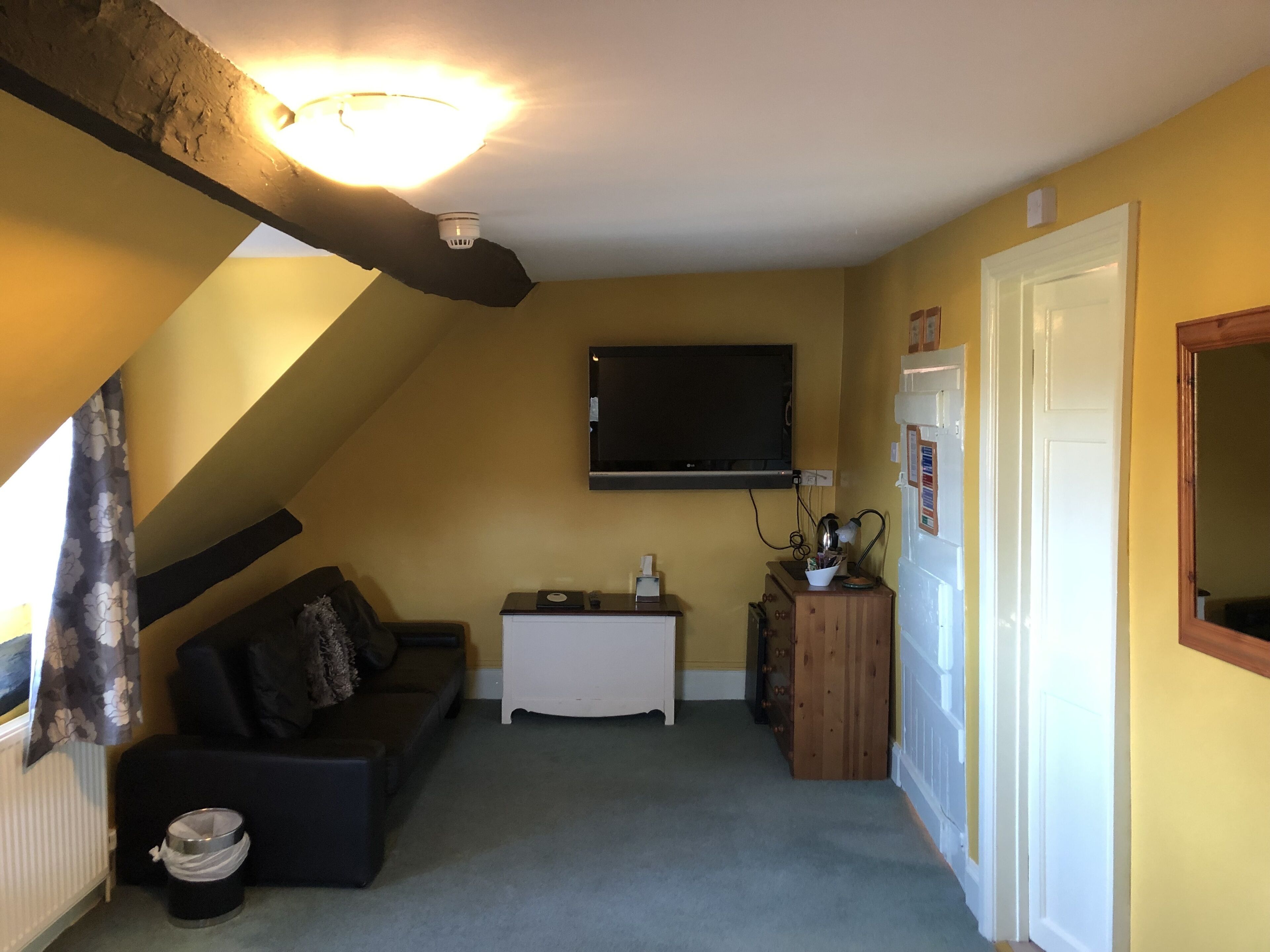 A compact living space with a sloped ceiling, a mounted TV, a small white cabinet, a wooden sideboard, and a black sofa with cushions.