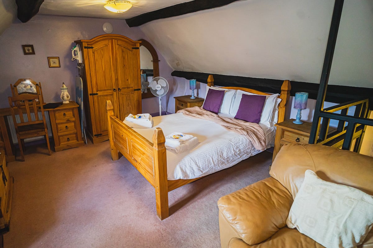 A bedroom with a wooden bed, a wooden wardrobe, a bedside table with a lamp, a chair, and a small table with a lamp.