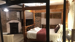 Ein Bett mit Baldachin, mit weißer Bettdecke und roter Decke, ein Holztisch mit weißem Tischtuch, ein Kamin aus Ziegelsteinen und ein Fernseher auf einem Schrank.