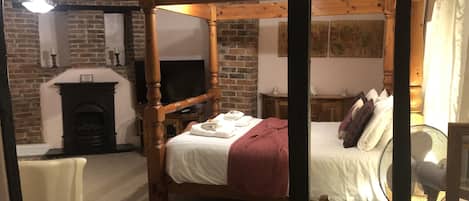 A four-poster bed with a white bedspread and red blanket, a wooden table with a white tablecloth, a brick fireplace, and a television on a stand.