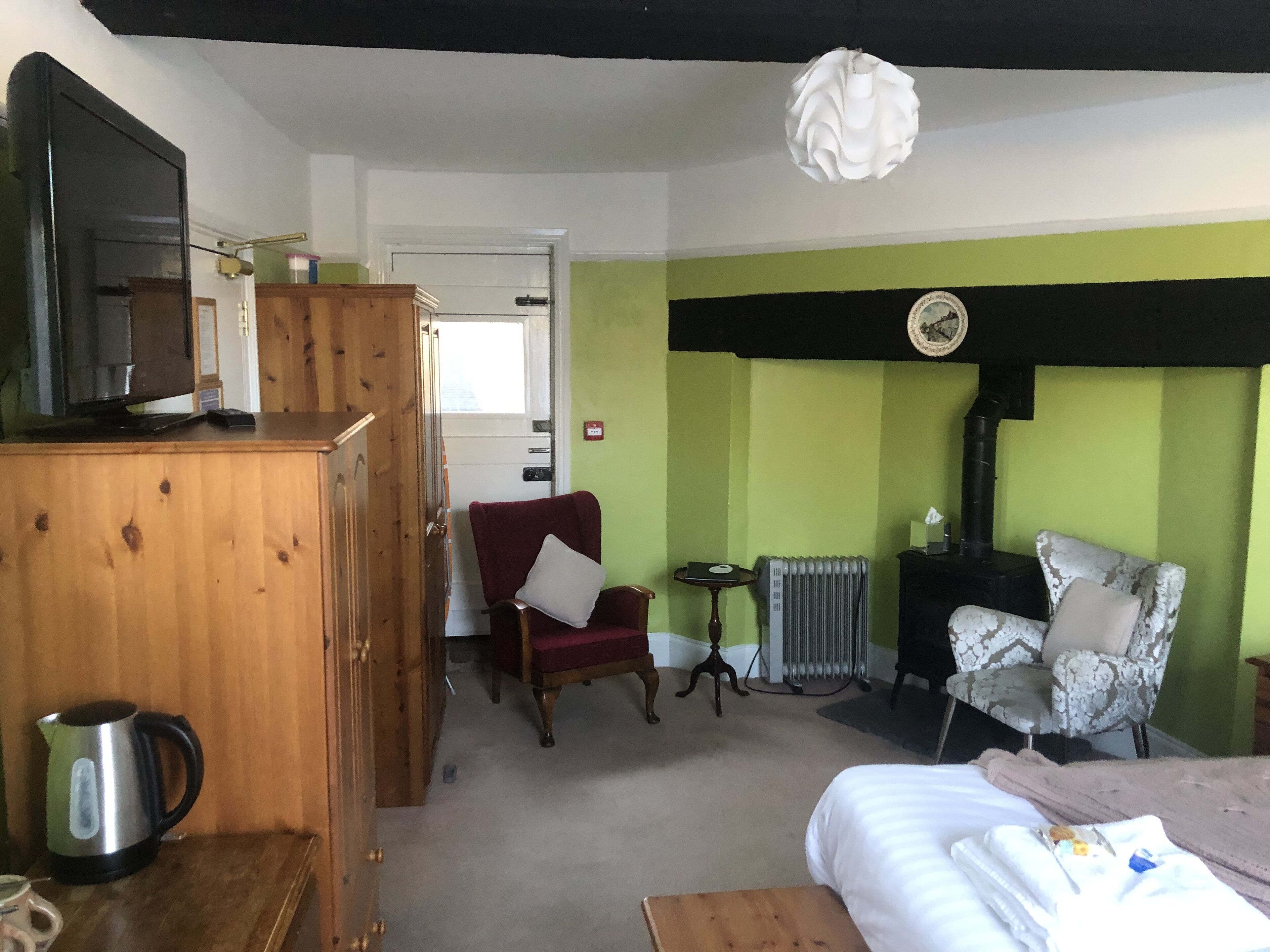 Una habitación con una cama, un aparador de madera, un sillón rojo, una silla blanca, un radiador y un televisor sobre un mueble de madera.