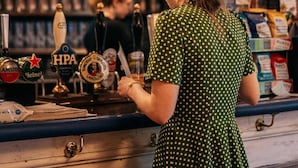 Eine Bar mit einer Frau im Punktekleid hinter der Theke, Zapfhähnen und Regalen mit Flaschen.