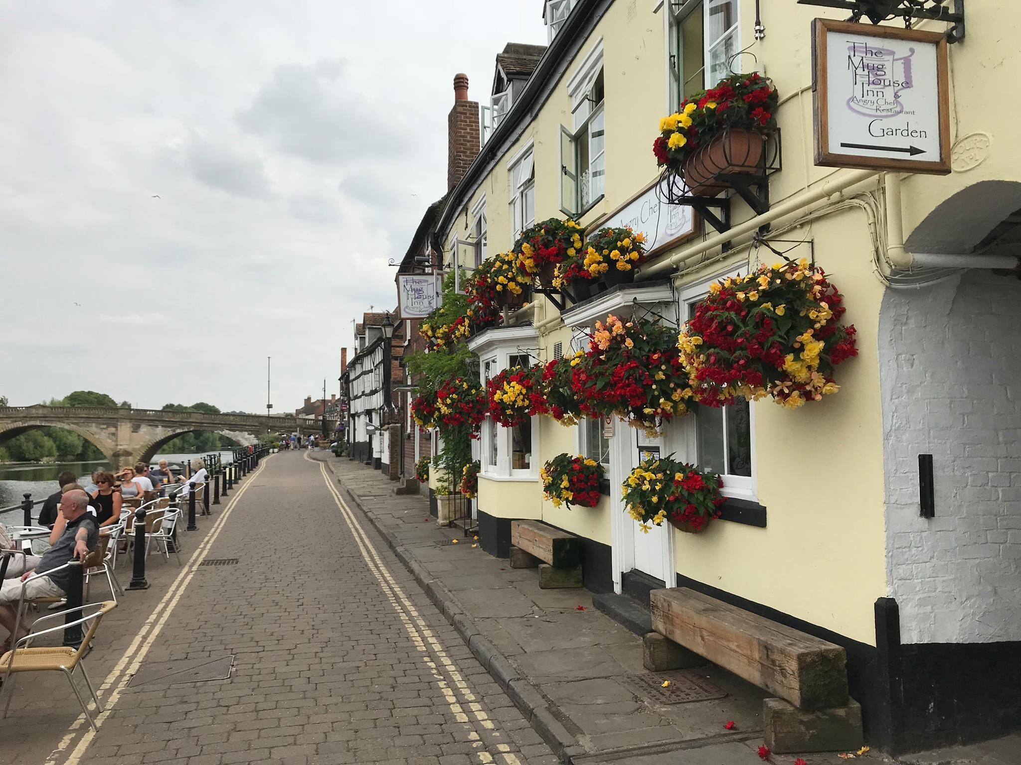 En rad traditionella byggnader med blomkorgar hängande från fönstren, en skylt som visar namnet på värdshuset "The Mug House Inn", och i bakgrunden syns en flod.