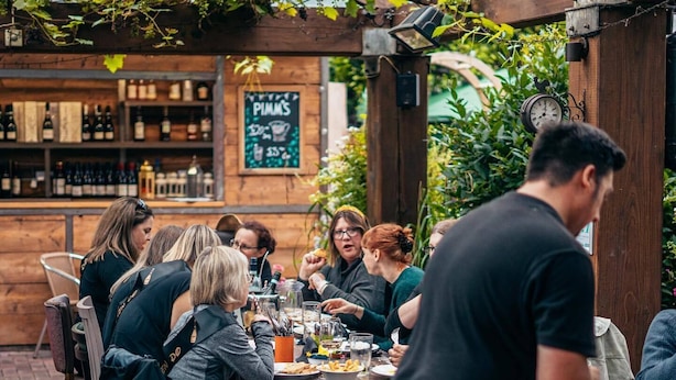 Eine Gruppe von Personen speist im Freien in einem Restaurant mit einer Holzpergola und einem Backsteinboden.