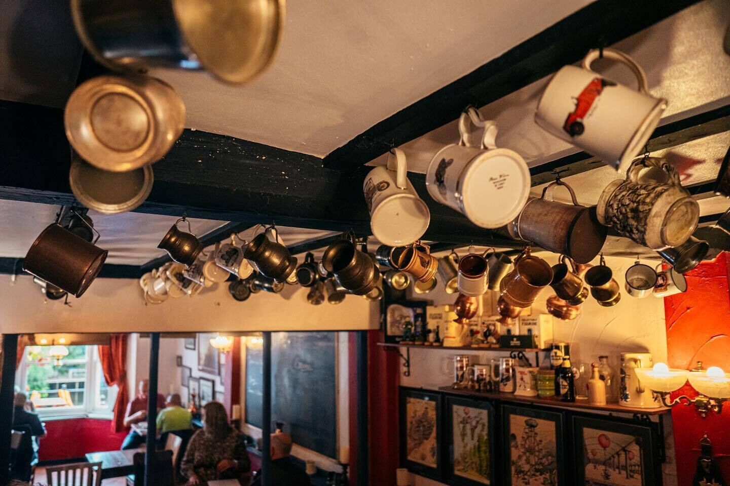 Um quarto com teto repleto de panelas e frigideiras penduradas, um espaço de bar com diversos frascos, e vista para a rua com pessoas lá fora.