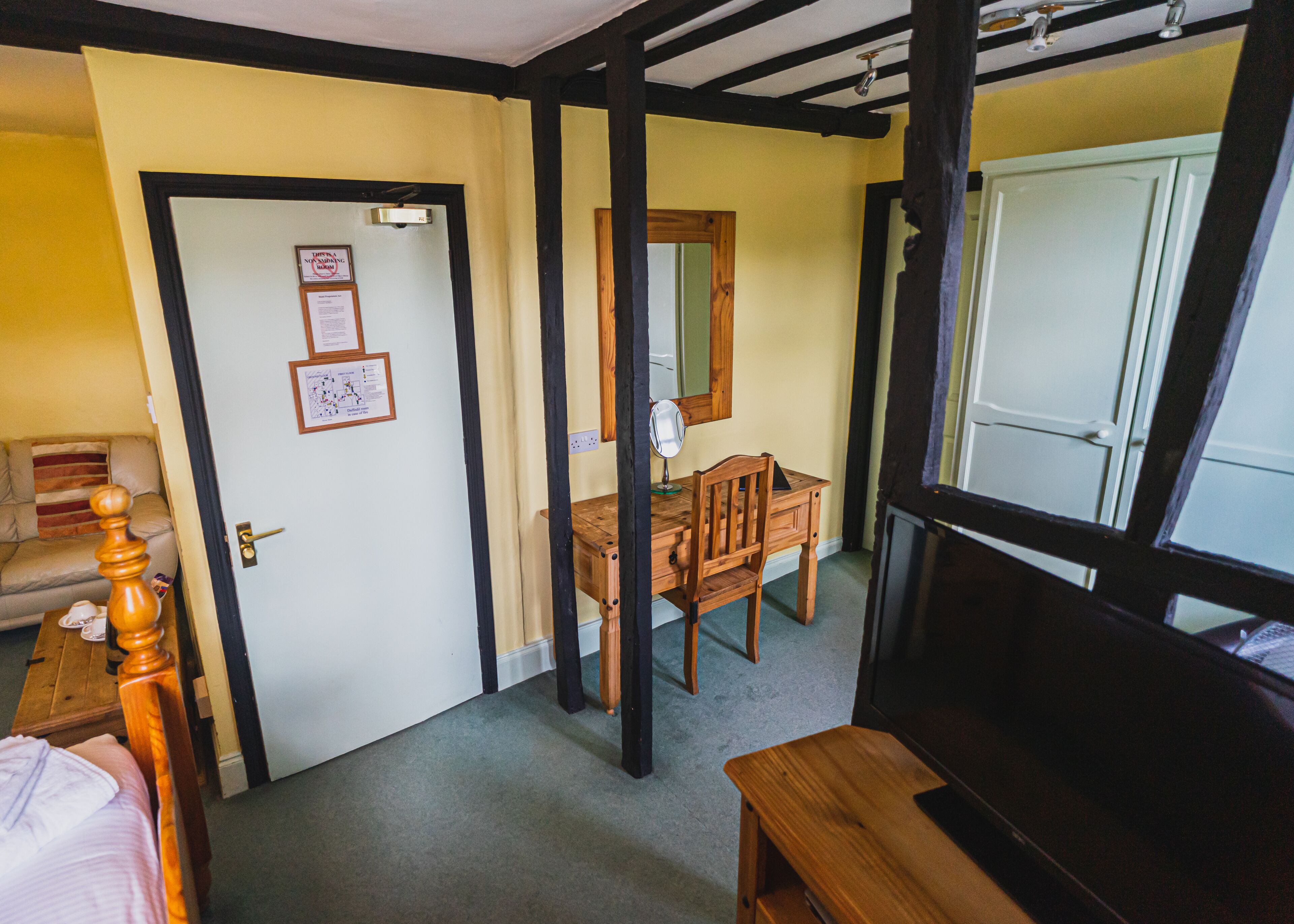 A room with a wooden desk, chair, and a mirror. There is a door with a sign on it, a bed with a wooden headboard, and a window with a view of another room.