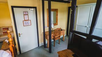 A room with a wooden desk, chair, and a mirror. There is a door with a sign on it, a bed with a wooden headboard, and a window with a view of another room.