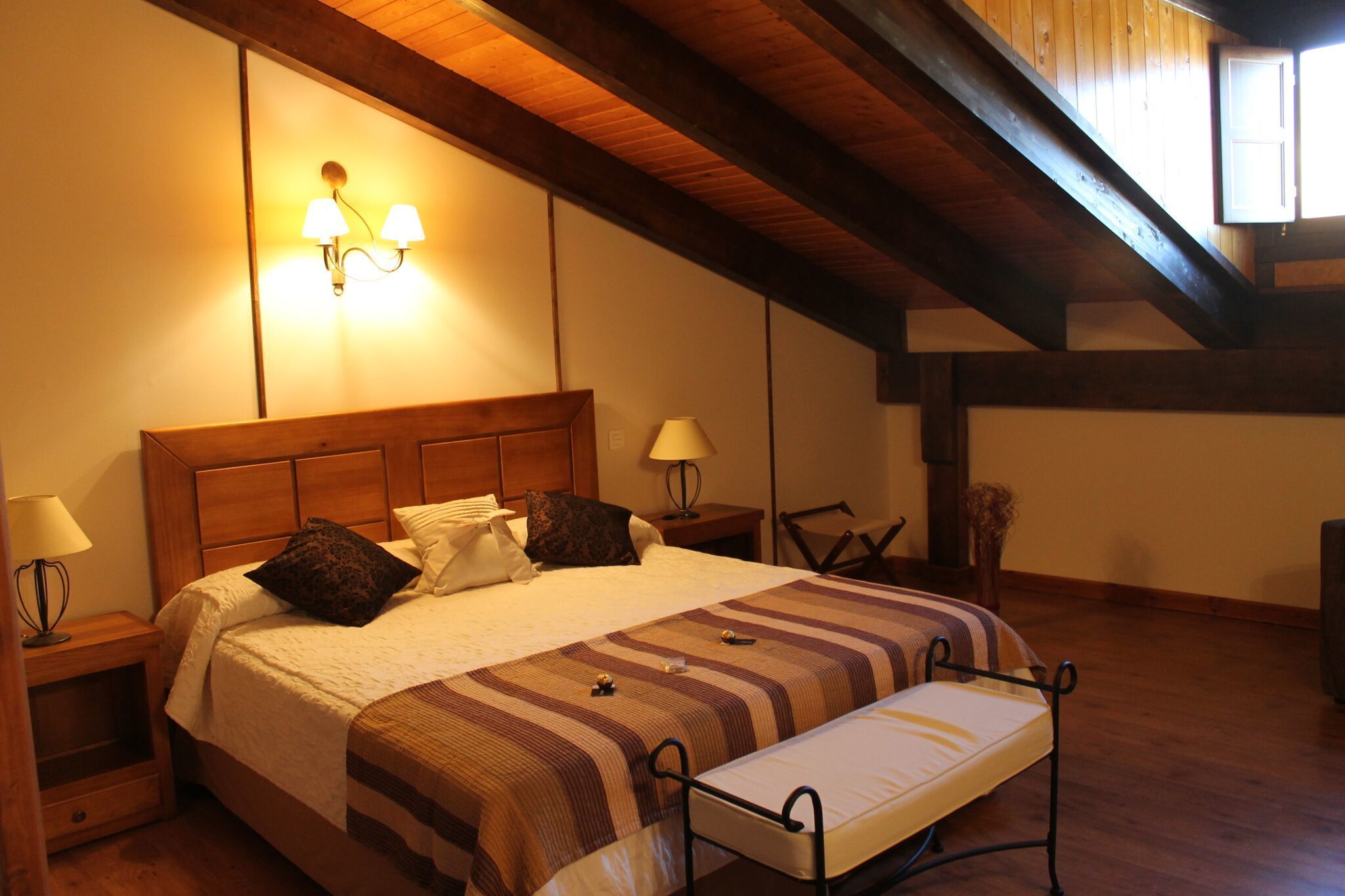 Un dormitorio con cabecera de madera, una cama grande con ropa de cama a rayas, dos lámparas de noche, un banco al pie de la cama y un techo inclinado con vigas a la vista.