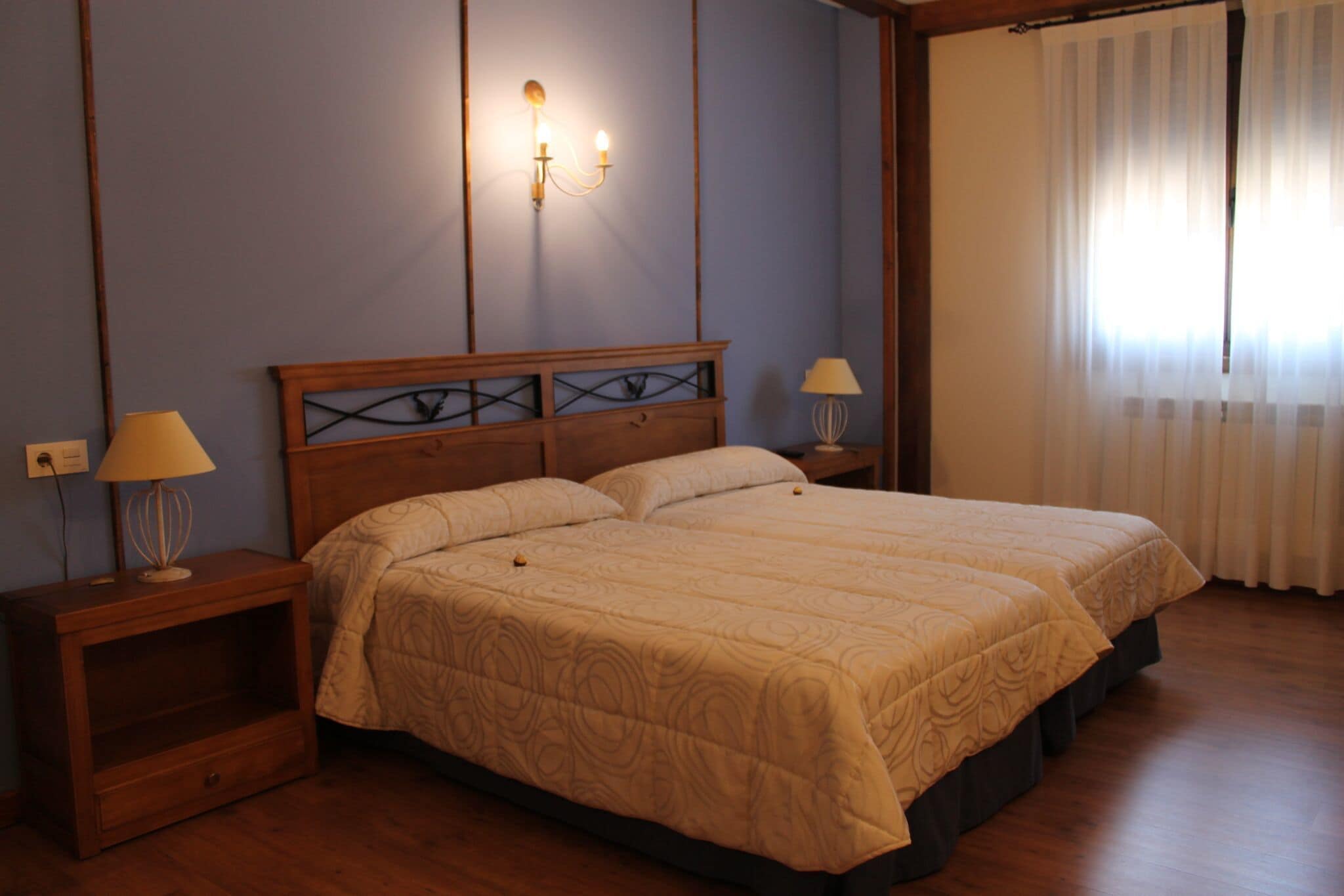 A double bed with a wooden headboard, two bedside tables with lamps, a nightstand, and a window with sheer curtains.
