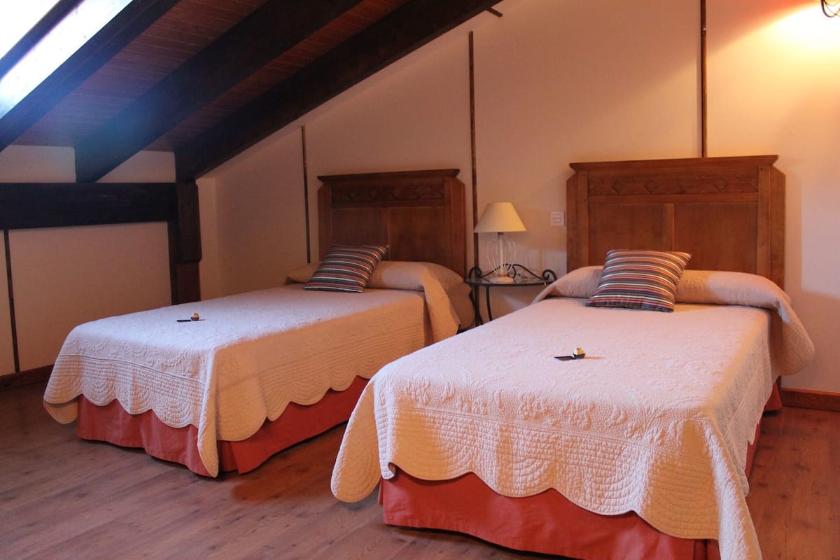 Deux lits simples avec des têtes de lit en bois et des couvre-lits rouges, une table de chevet avec une lampe, et un plafond en pente avec des poutres apparentes.