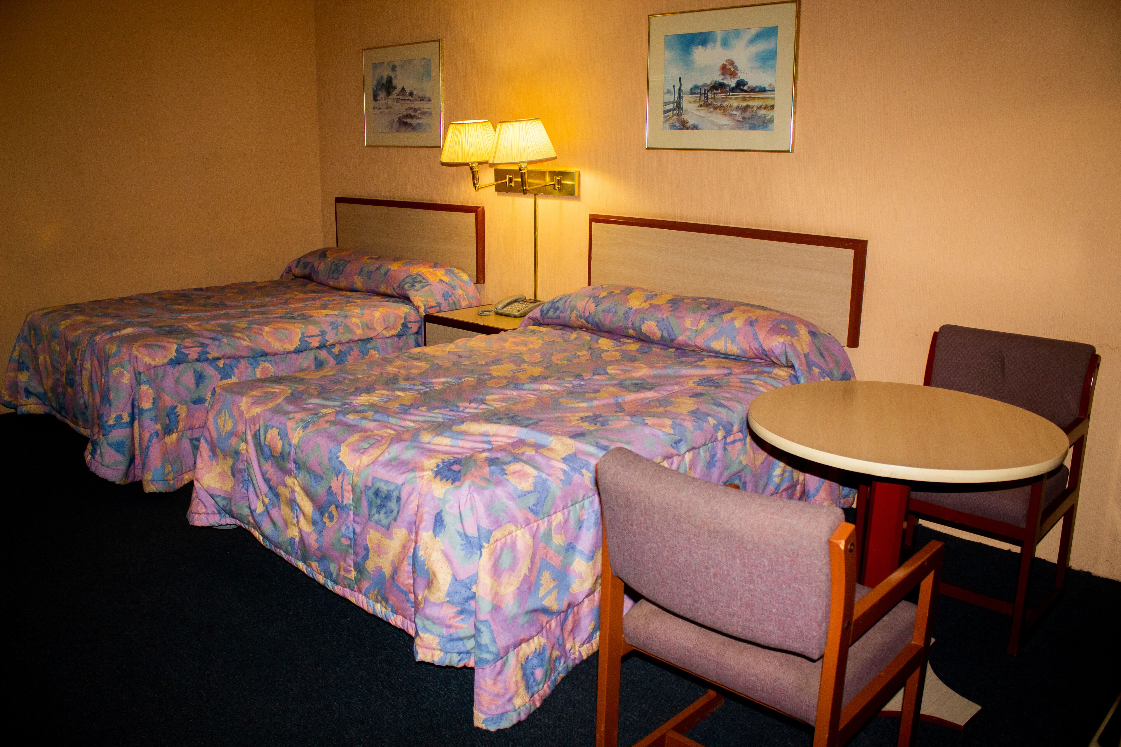 Habitación de hotel con dos camas, una mesita y una silla. Hay dos lámparas de pared y dos cuadros enmarcados.