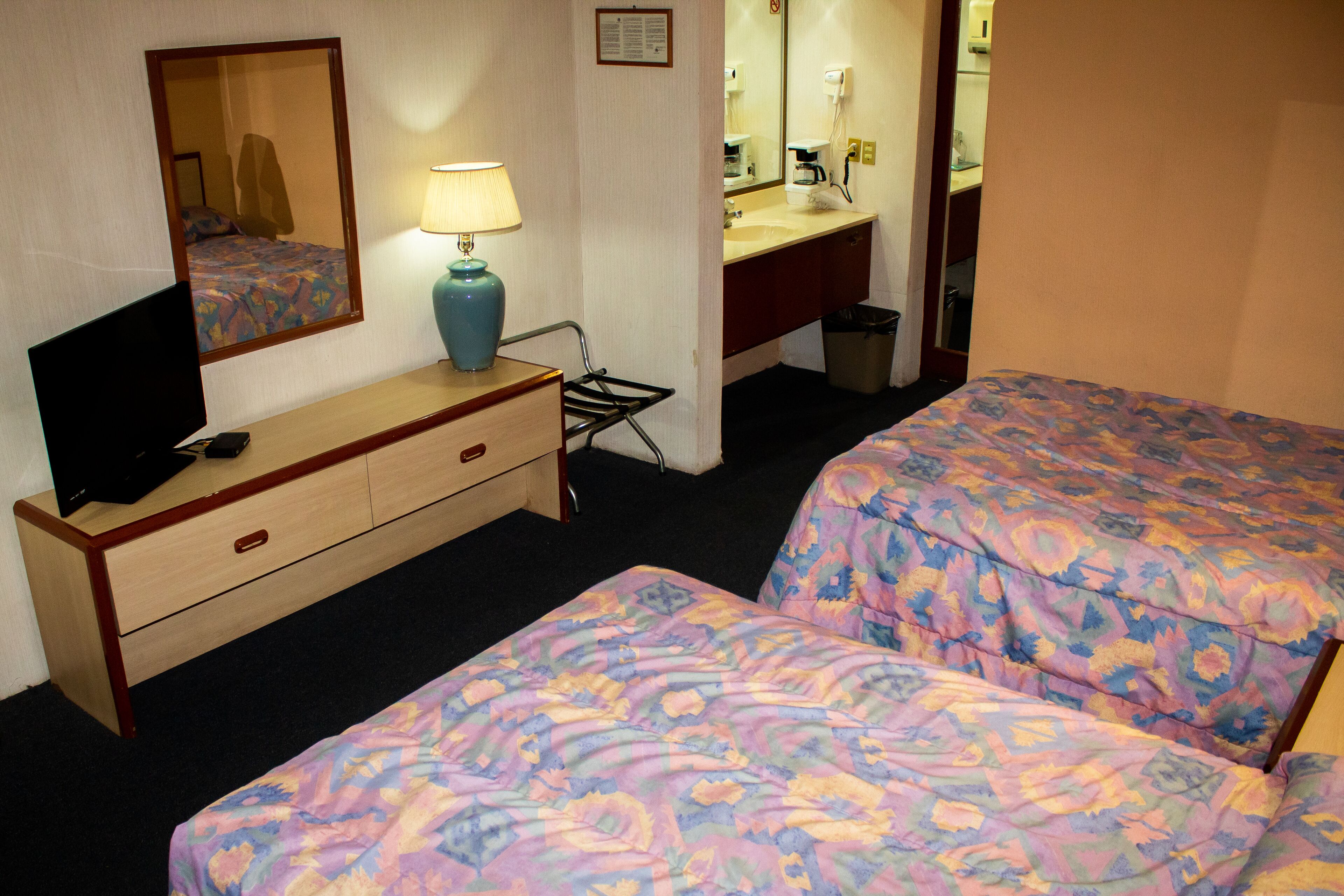 Habitación de hotel con dos camas, un escritorio con televisor, una lámpara, un espejo, un baño con lavamanos y espejo, y un cesto de basura.