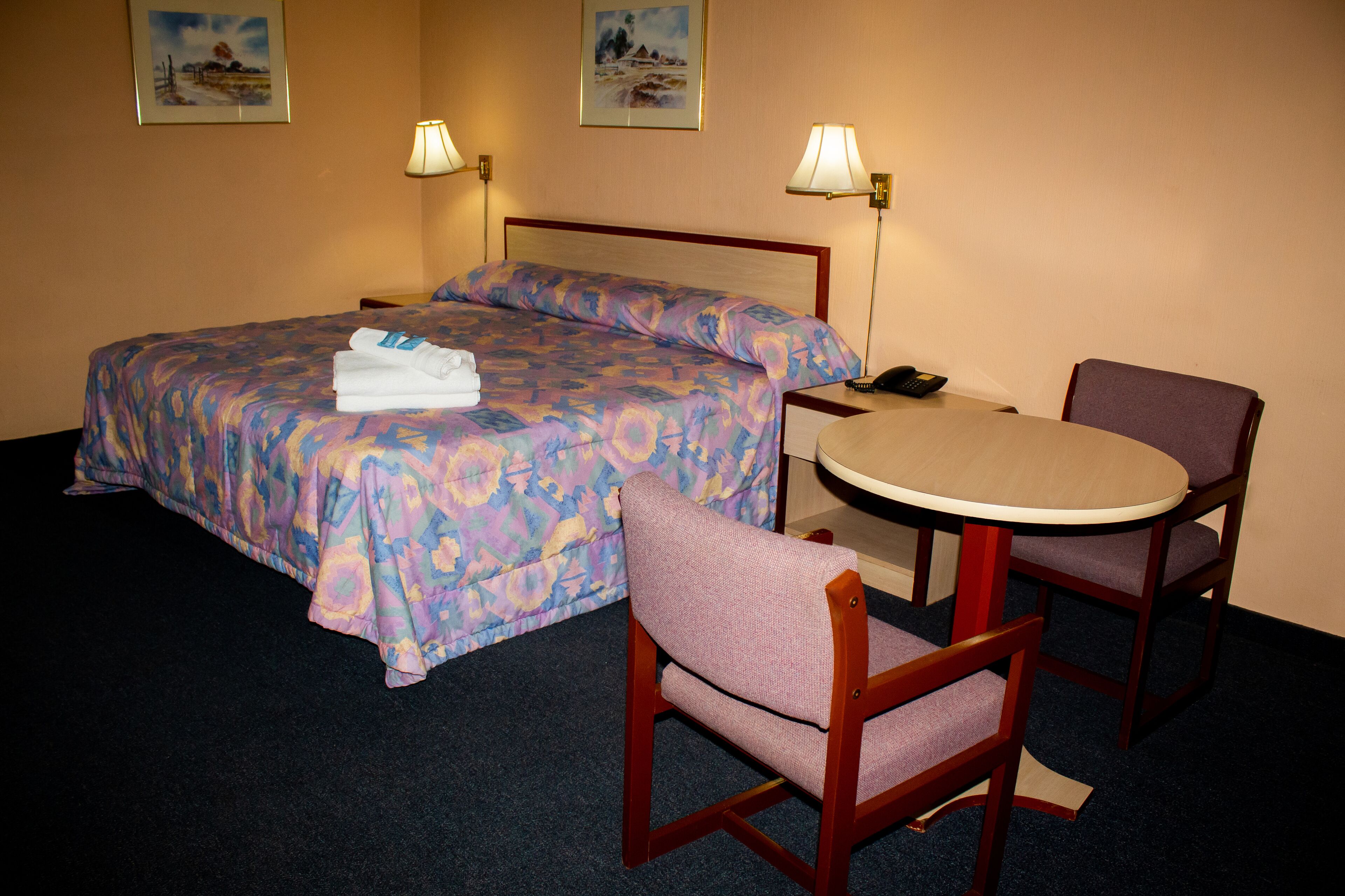 Una habitación de hotel con una cama, dos sillas, una mesita y dos lámparas de pared.