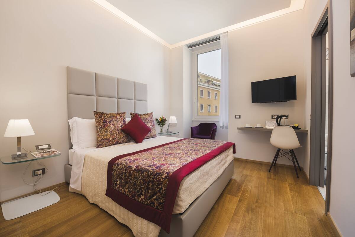 Une chambre moderne avec un grand lit, une télévision et une fenêtre donnant sur des immeubles.