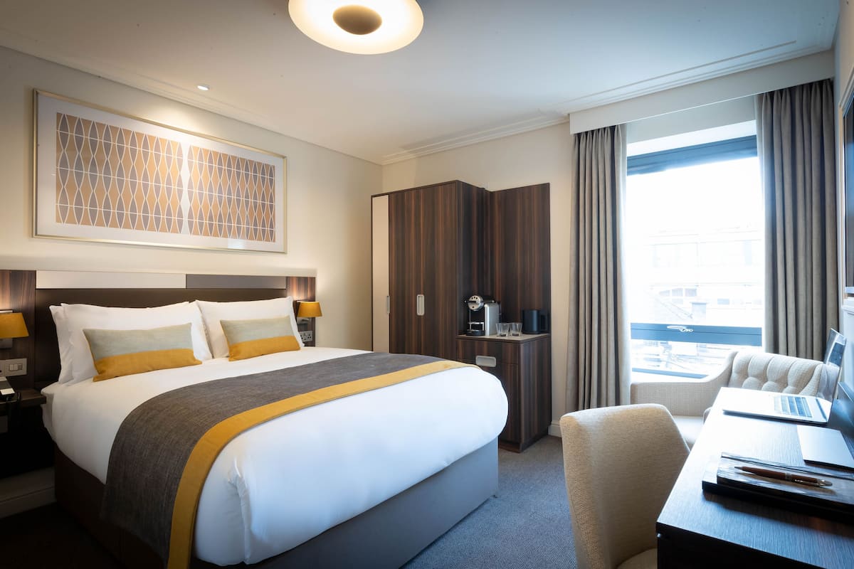 Habitación de hotel con una cama grande, un escritorio con una computadora portátil, un mini bar y vistas a la ciudad a través de amplios ventanales.